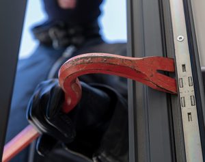 Header-Artikel: Ein Dieb mit Sturmhaube hält ein Brecheisen in der Hand, um ein Glasfenster einzuschlagen.