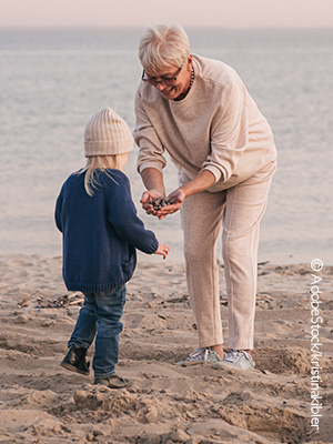 Eine ältere Dame ist mit einem Kind am Strand und zeigt ihm Steine. 