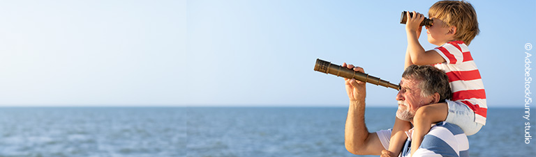 Ein Junge sitzt auf den Schultern seines Opas. Gemeinsam schauen sie mit Ferngläsern zum Meer.
