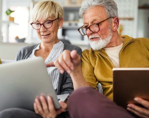 Eine ältere Frau und ein Mann sitzen auf dem Sofa im Wohnzimmer und schauen in ihre Tablets.