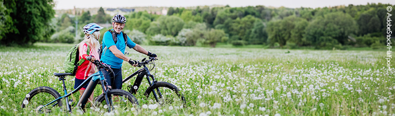 Eine Mann und eine Frau stehen mit ihren E-Bikes in einer blühenden Blumenwiese und lächeln sich an. 